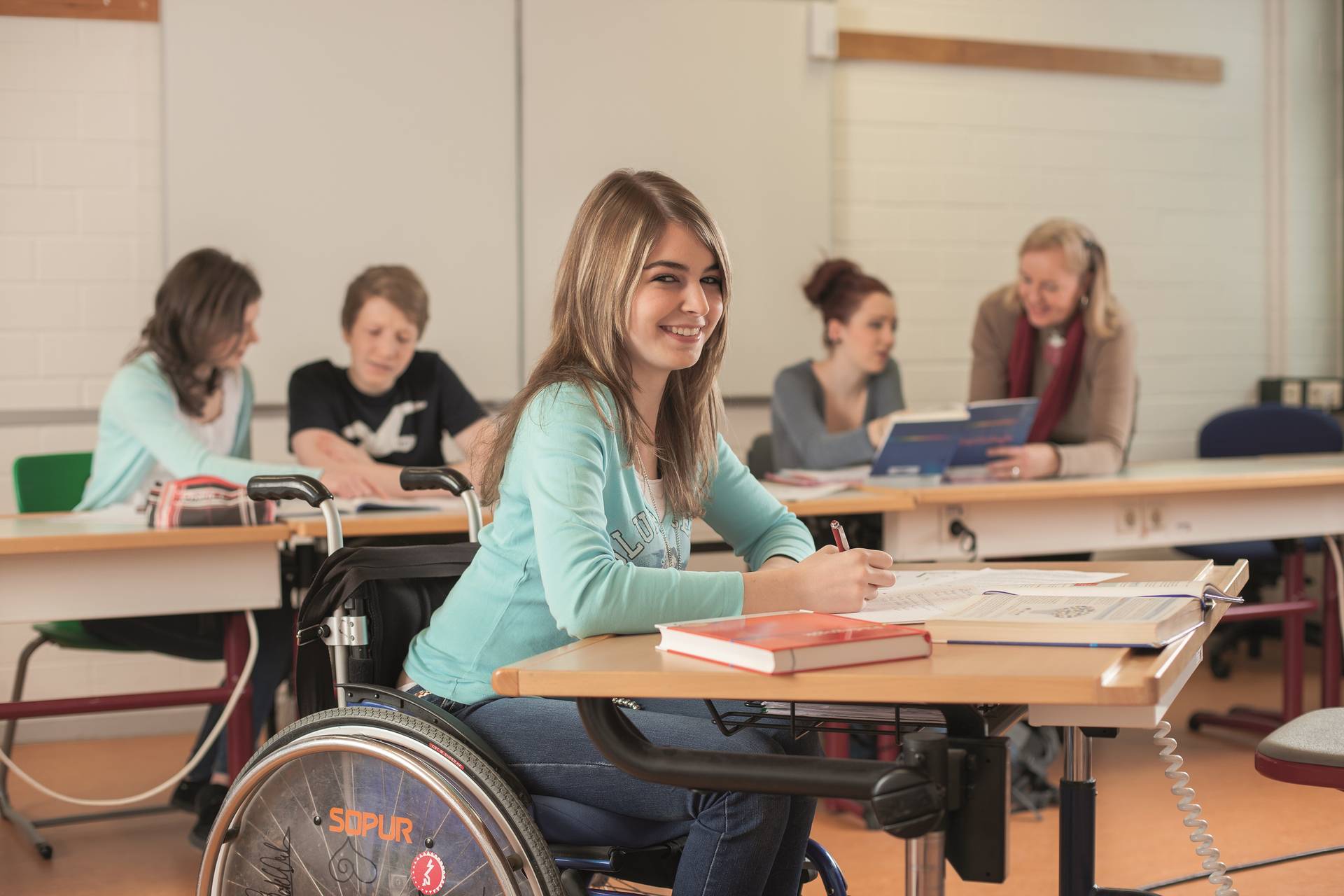 Schülerin im Rollstuhl lächelt während des Unterrichtsgeschehens in die Kamera.