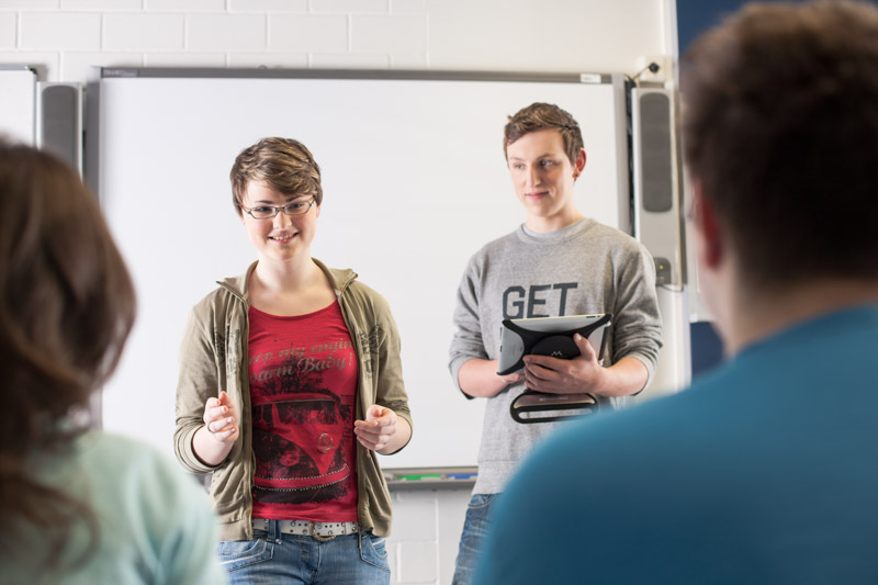 Schülerin hält ein Referat. Ein Schüler unterstützt mit einem Tablet die Präsentation.