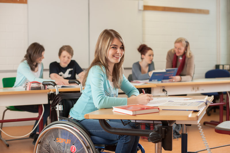 Schülerin im Rollstuhl lächelt während des Unterrichtsgeschehens in die Kamera.