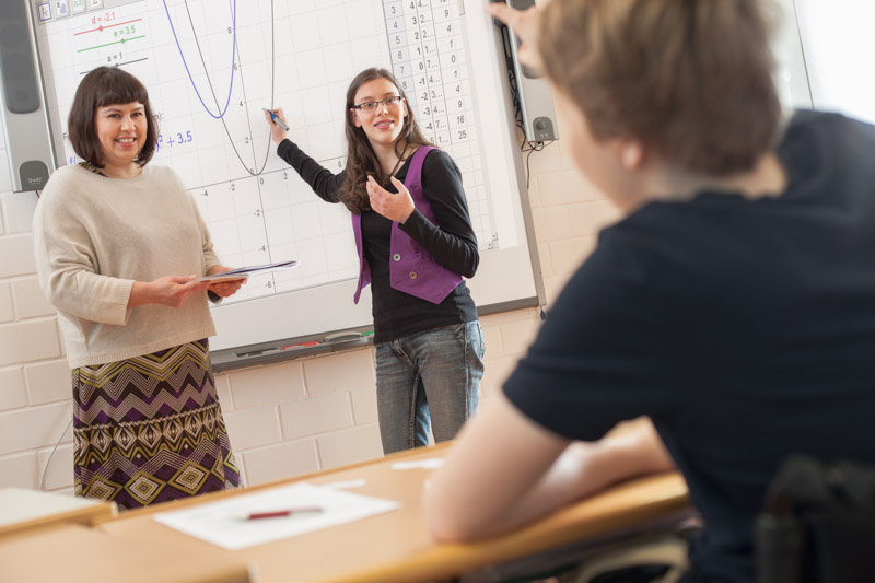 Schülerin und Lehrerin stehen vor der Klasse und erklären ein digitales Schaubild.