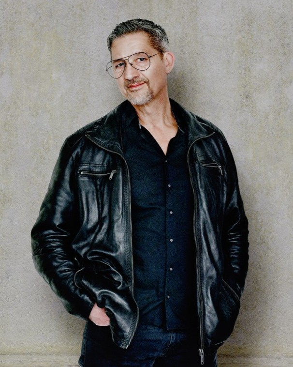 Musiker Robert Matt steht vor einer Betonwand. Er trägt ein dunkles Hemd und eine schwarze Jacke.