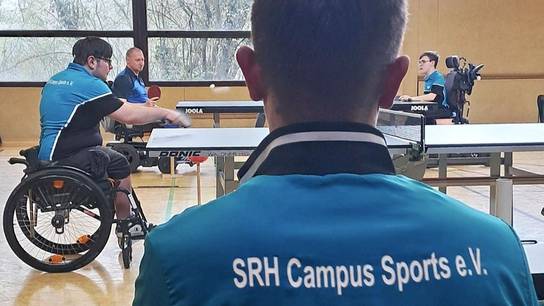 Das SRH-SHS-Rollstuhl-Tischtennis-Team beim Tischtennisspiel; im Vordergrund junger Mann mit SRH-Campus-Sport-T-Shirt