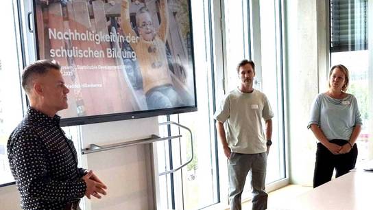 Drei Personen in einem Raum mit Präsentationen auf Bildschirmen.