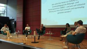 Politiker:innen und zwei Schüler-Moderatoren sitzen im Rahmen der Podiumsdiskussion zum Thema Demokratie und Inklusion im Fokus auf der Aula-Bühne. Im Hintergrund wird Text auf eine leinwand projiziert.