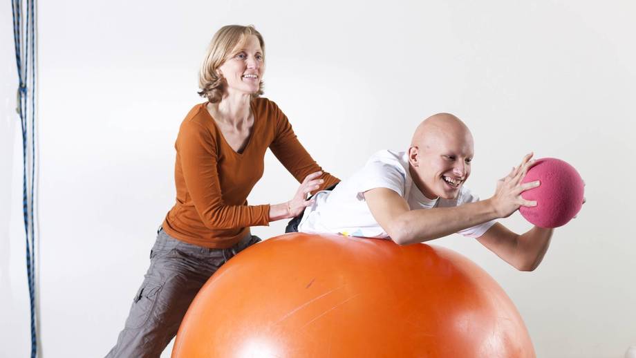 Schüler übt eine physiotherapeutische Übung am Gymnastikball aus. Die Physiotherapeutin unterstützt ihn dabei.
