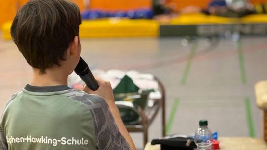 Schüler moderiert die Tischtennis-Übernachtung; auf dem T-Shirt (Rücken) ist der Schulname SRH Stephen-Hawking-Schule aufgedruckt.