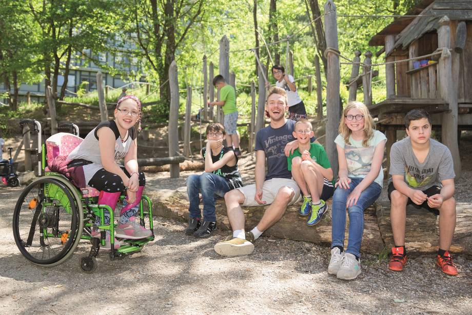 Schüler:innen und ein Erzieher sitzen nebeneinander auf einem liegenden Baumstamm.