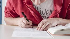 Schüler schreibt fleißig auf ein Blatt Papier. Daneben liegt ein aufgeschlagenes Lehrbuch.