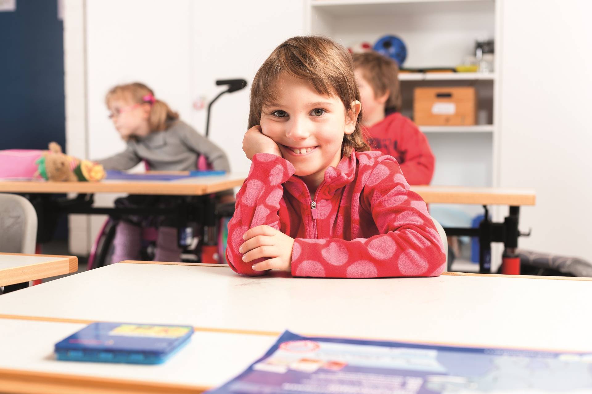 Grundschülerin sitzt hinter der Schulbank und grinst in die Kamera.