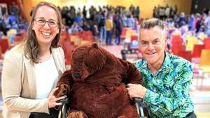 Zwei Lehrkräfte der SEH Stephen-Hawking-Schule lachen in die Kamera. Zwischen ihnen sitzt ein großer Teddybär im Rollstuhl.