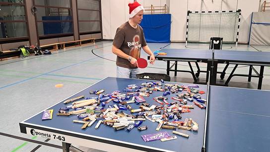 Zahlreiche Süßigkeiten liegen auf einer Tischtennisplatte in der Turnhalle; der Trainer befindet sich mit Weihnachtsmannmütze im Hintergrund