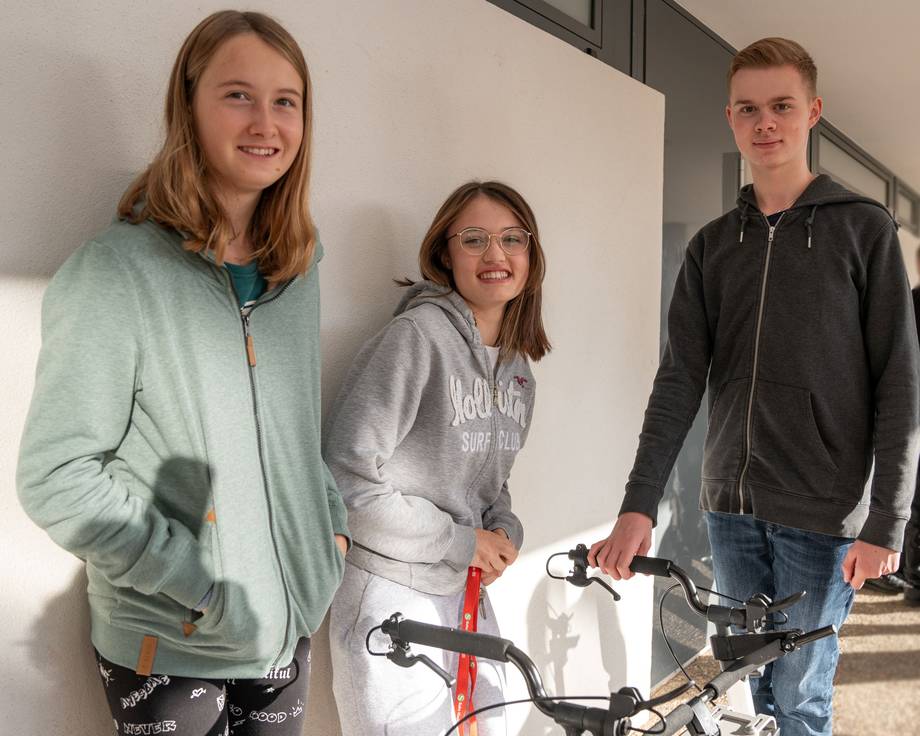 Drei Schüler:innen stehen in der großen Hofpause auf dem Schulhof und lachen in die Kamera.