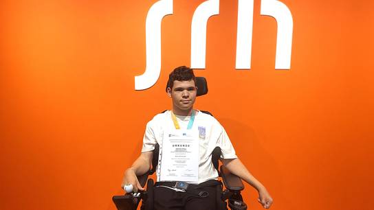 Schüler Johannes zeigt die Urkunde der erfolgreichen Teilnahme; im Hintergrund ist das SRH-Logo zu sehen