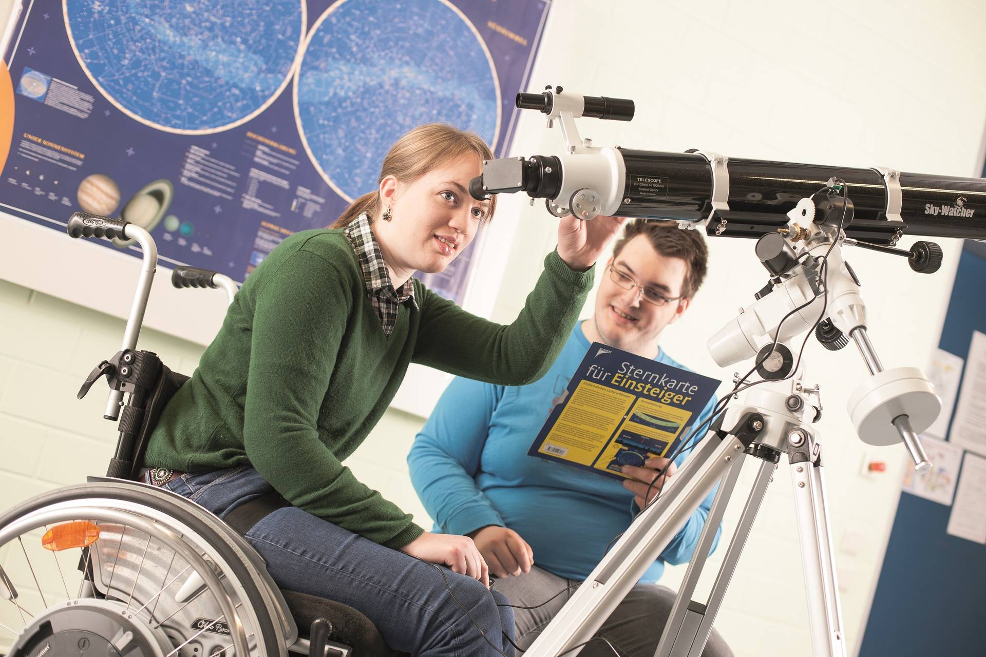 Schülerin im Rollstuhl schaut durch ein Teleskop. Daneben sitzt ein Schüler mit Sternenkarte in der Hand.
