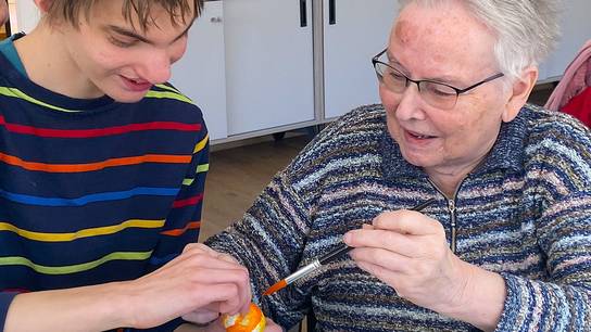 Schüler bemalt gemeinsam mit einer Seniorin mithilfe eines Pinsels ein Osterei.