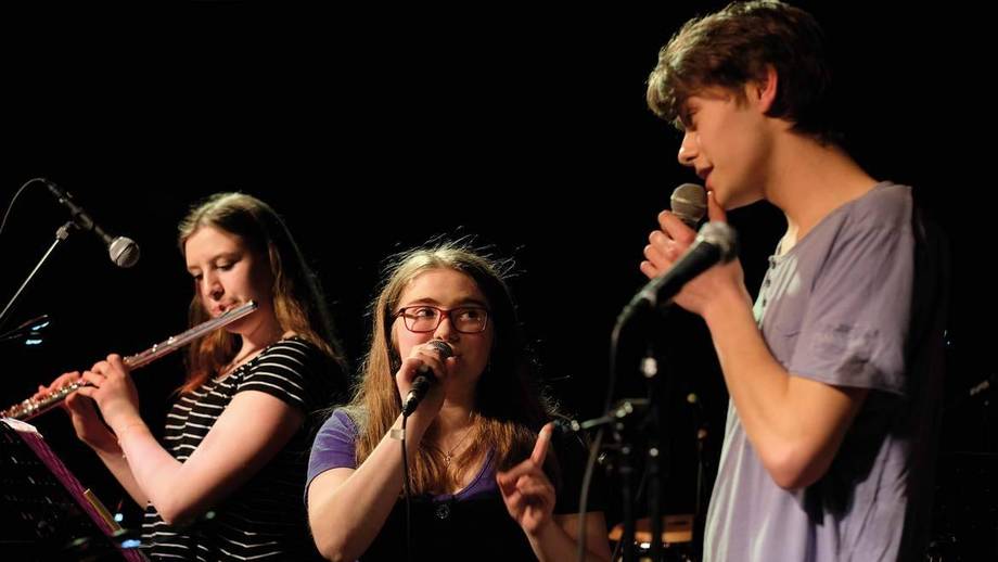 Zwei Schülerinnen und ein Schüler geben ein Schülerbandkonzert. Sie singen und spielen Querflöte.